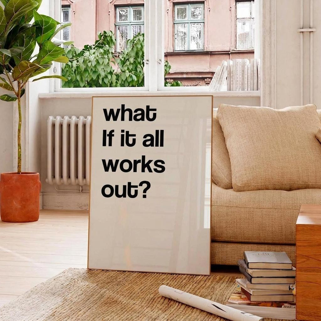 A vertical framed art print with the text 'what If it all works out?' in black sans-serif font on a white background, leaning against a beige sofa in a bright, modern living room.