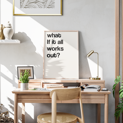 A framed typographic wall art print with the text 'what if it all works out?' displayed on a light wood desk in a styled home office setting with a wooden chair, lamp, and plants.