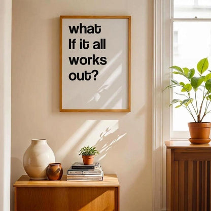 A framed typographic wall print reading "what if it all works out?" hangs on a beige wall above a wooden side table in a sunlit room with a window, potted plant, and decorative vases.